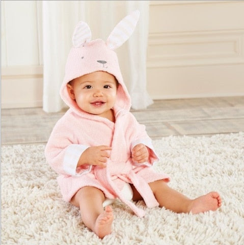 Lille barn i lyserød kaninbadekåbe sidder smilende på hvidt tæppe i stue. Sødt babyoutfit, kaninører, hyggelig indretning.