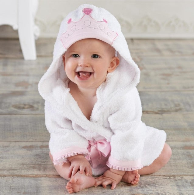 Baby iført hvid badekåbe med hætte og lyserød krone, sidder smilende på trægulv. Sødt babytøj, badetid, baby mode.