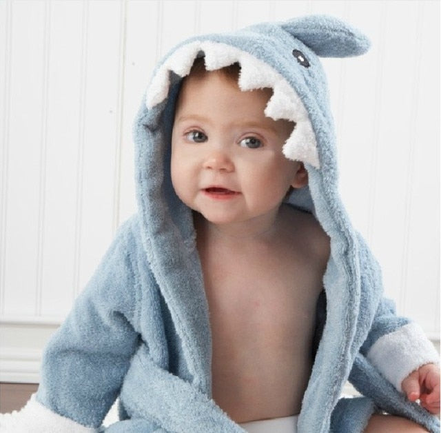 Baby iført blå hajbadekåbe med hætte, sidder foran hvid panelvæg. Sødt barn i sjovt tøj, baby mode, haj tema, indendørs portræt.