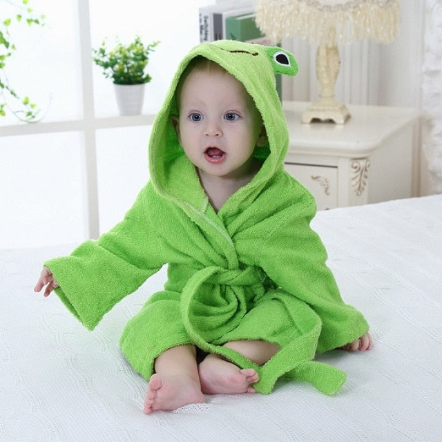 Baby iført grøn frø-badekåbe sidder på seng. Sødt barn i blødt, dyreinspireret tøj. Perfekt tilbehør til babybadetid.