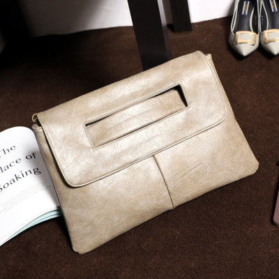 Beige læder clutch taske på brun baggrund ved siden af åbnet bog og grå højhælede sko. Elegant design, praktisk til modebevidste kvinder.