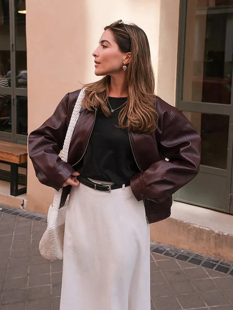 Kvinde i brun læderjakke og hvid nederdel, poserer på gaden. Hun bærer en hvid nettaske og solbriller i håret. Mode, street style, efterår.