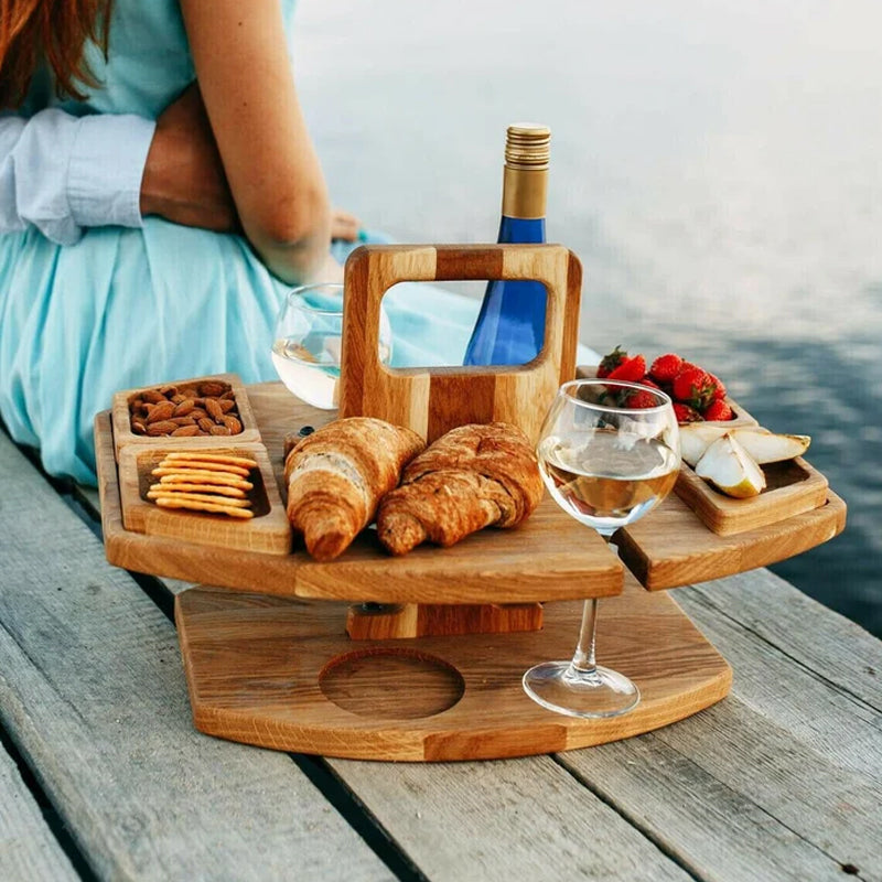 Picnicbord i træ med croissanter, ost, jordbær, mandler, kiks, vinflaske og vinglas. Perfekt til romantisk udendørs picnic ved vandet.