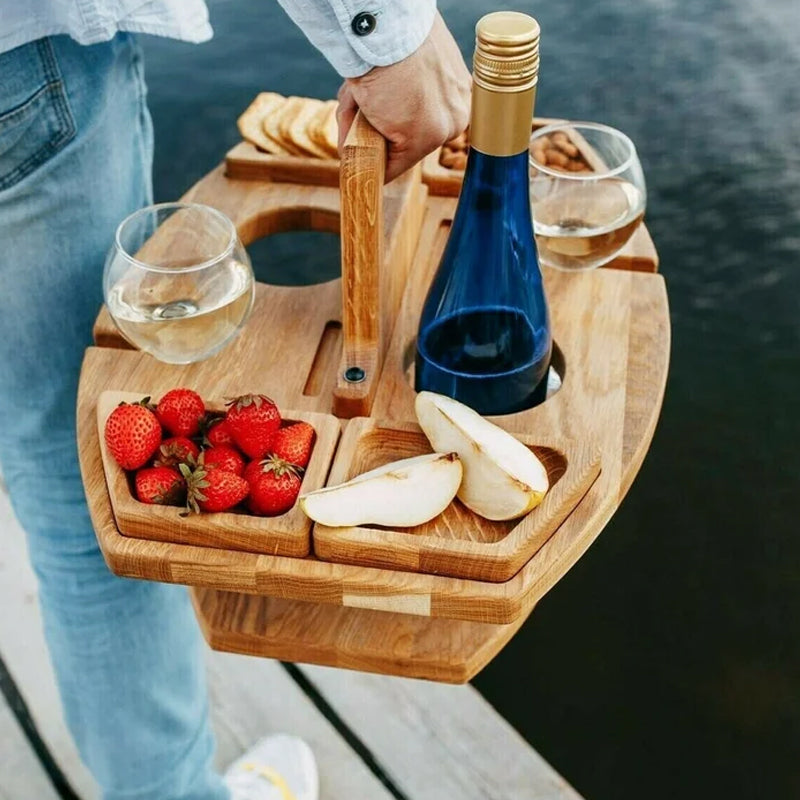 Person bærer en træbakke med vinflaske, to glas, jordbær, pærer og snacks ved en sø. Perfekt til picnic, udendørs hygge og romantiske stunder.