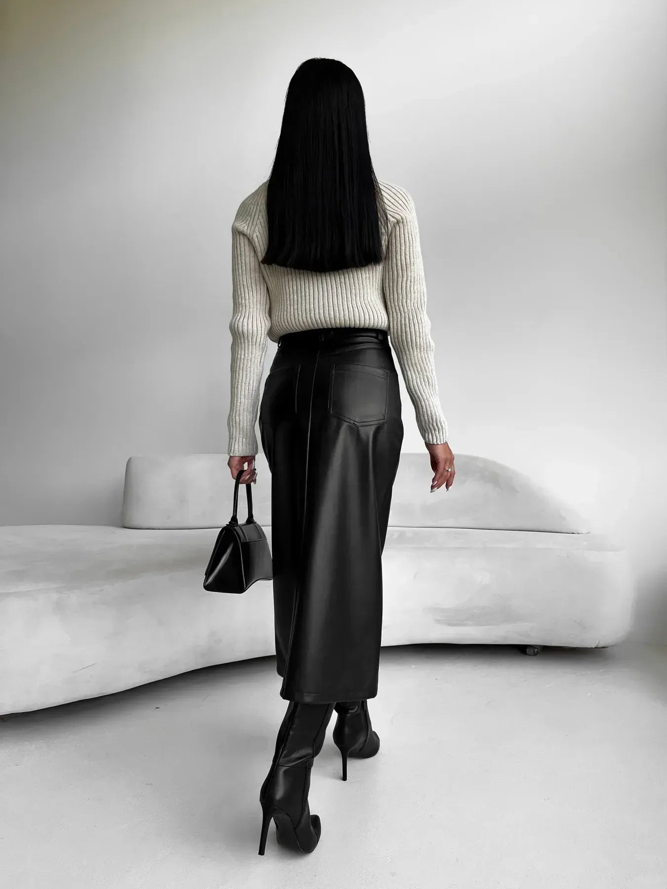Kvinde i hvid sweater og sort lædernederdel, bærer sort taske og høje hæle, står foran hvid sofa. Mode, stilfuldt outfit, minimalistisk indretning.