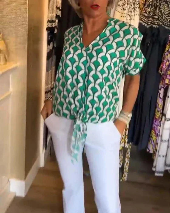 Kvinde iført grøn og hvid mønstret bluse med knude, hvide bukser, står i tøjbutik. Moderne damemode, sommeroutfit, stilfuldt design.