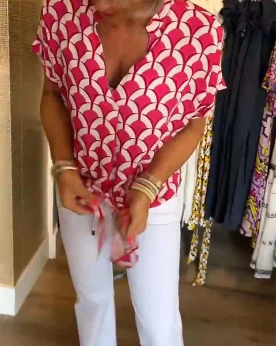 Kvinde iført rød-hvid mønstret bluse og hvide bukser, står i tøjbutik. Moderne mode, farverigt design, stilfuldt outfit, shopping.