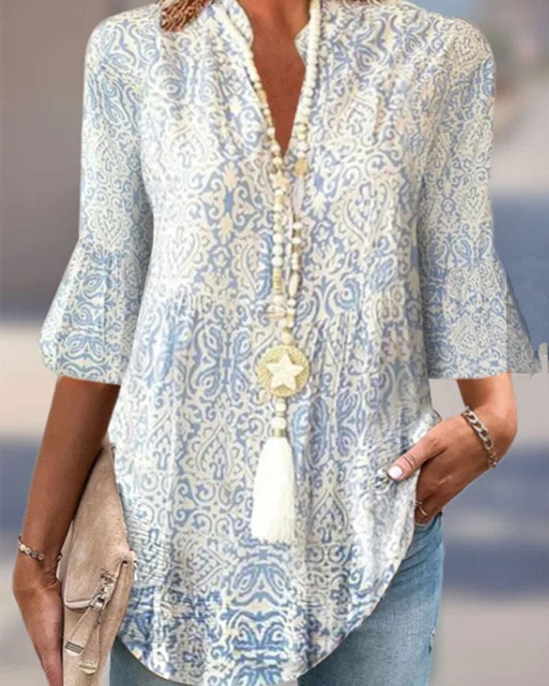 Dame i mønstret bluse med blå og hvide detaljer, iført halskæde med stjernevedhæng. Holder en beige taske. Casual og stilfuldt look.