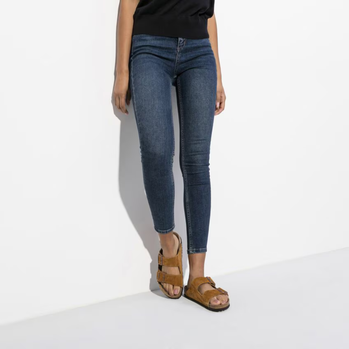 Kvinde iført mørkeblå skinny jeans og brune Birkenstock sandaler mod hvid baggrund. Mode, casual tøj, komfortabelt fodtøj.