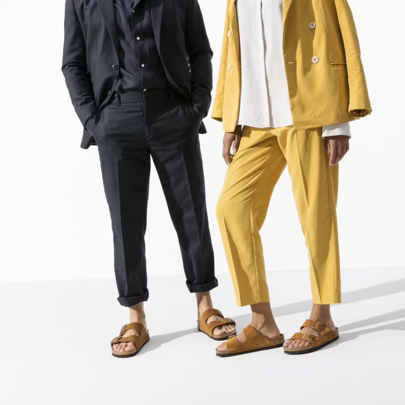 To personer iført moderne jakkesæt, en i mørkeblå og en i gul, med brune sandaler. Stilfuldt tøj, minimalistisk mode, trendy look.