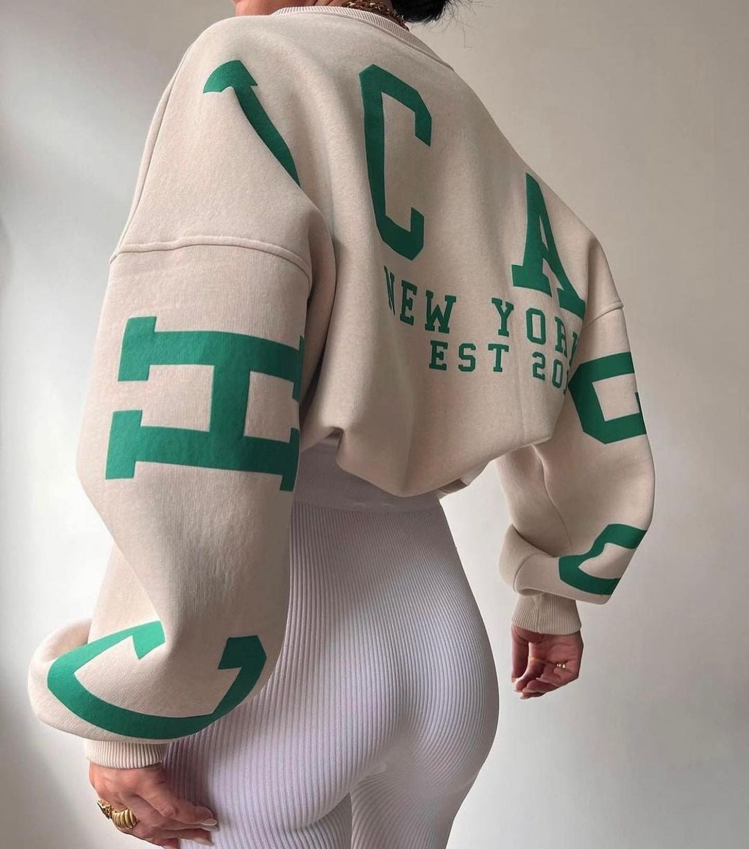 Beige sweatshirt med grønne bogstaver, "New York Est 2020", bæres af person i hvide ribbede leggings. Mode, streetwear, afslappet stil.