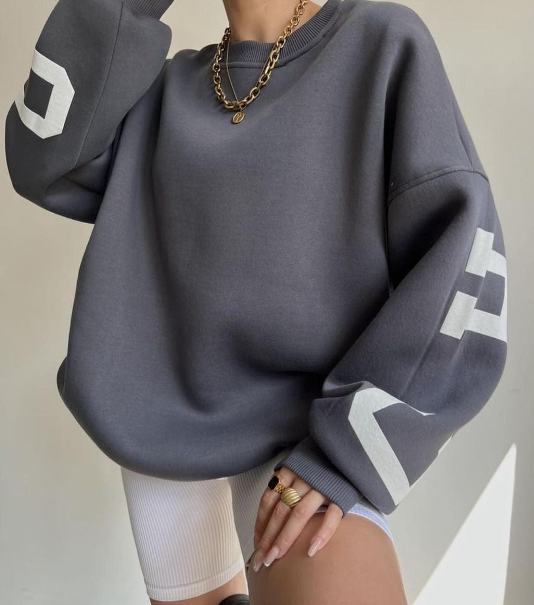 Kvinde iført grå oversized sweatshirt med hvide bogstaver på ærmerne, guldhalskæde og hvide cykelshorts. Trendy streetwear mode.