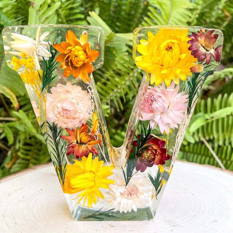 Klar harpiksbogstav "V" med indlejrede farverige blomster, placeret på en træskive foran grønne bregner. Unik dekoration, naturinspireret design.