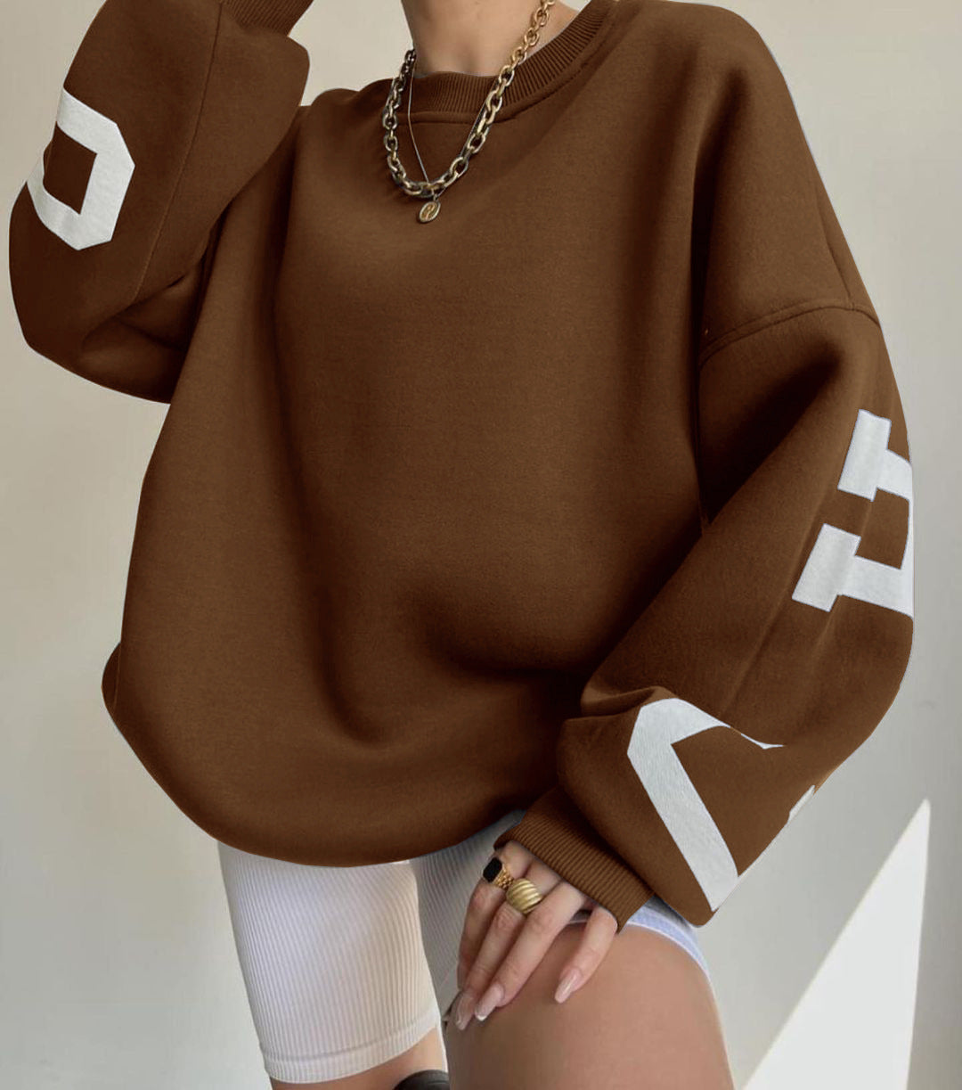 Brun sweatshirt med hvide bogstaver på ærmerne, båret af person med guldkæde og ringe, kombineret med hvide cykelshorts. Trendy streetwear look.