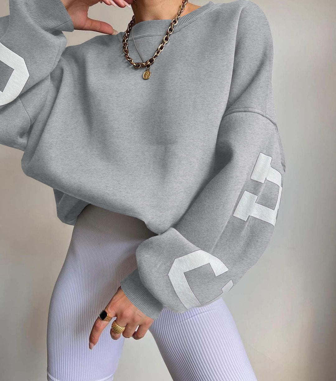 Grå oversized sweatshirt med hvide bogstaver, båret af person med guldkæde og ringe, kombineret med hvide ribbede leggings. Trendy streetwear look.