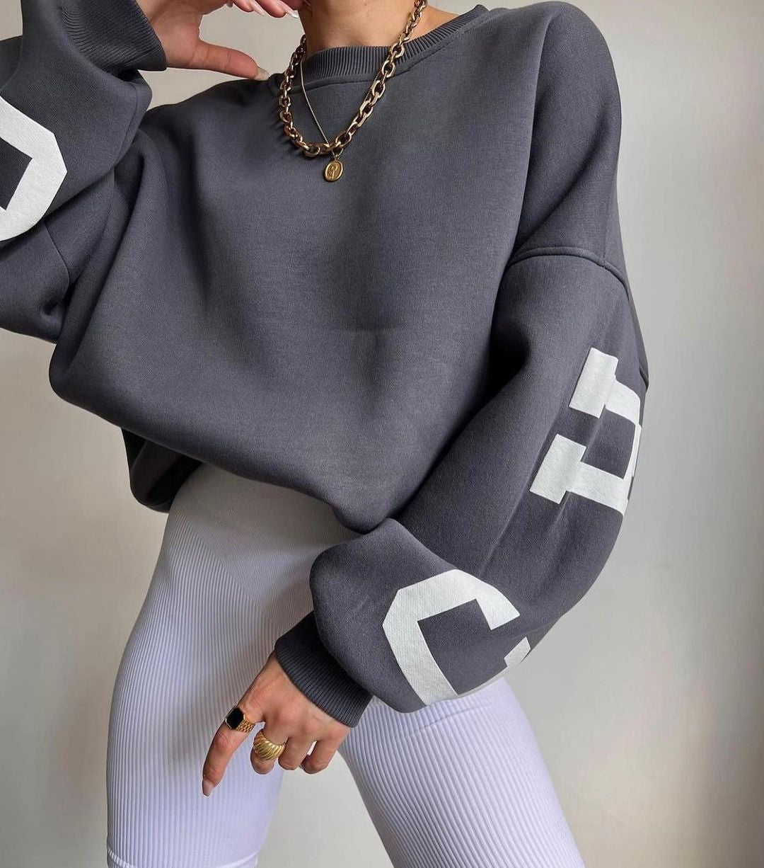 Grå oversized sweatshirt med hvide bogstaver, båret af person med hvid ribbet leggings og guldkæde. Trendy streetwear mode.