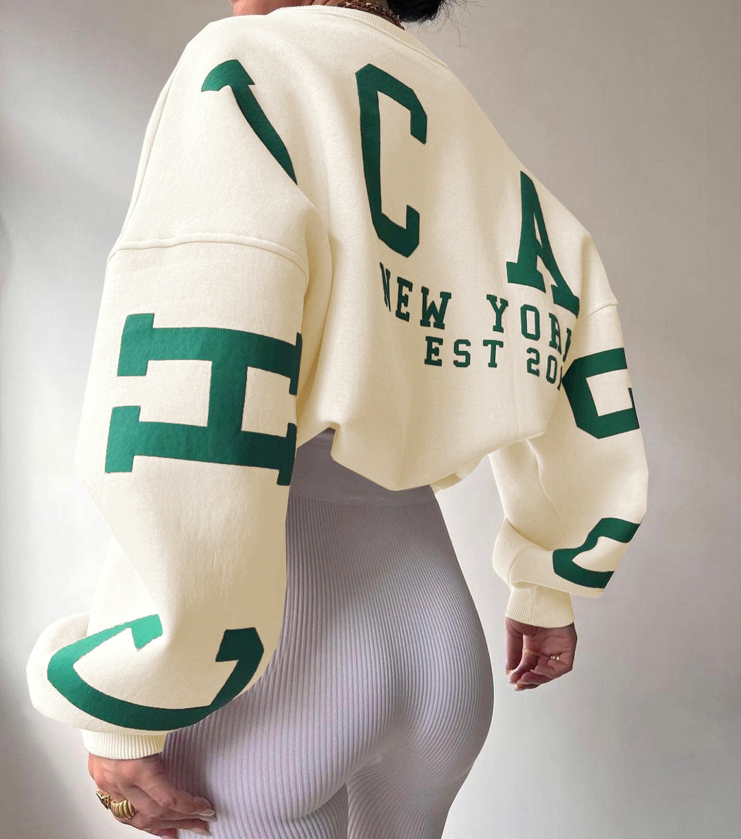 Dame i hvid sweatshirt med grønne bogstaver, "NEW YORK EST 2020" på ryggen. Moderne streetwear, afslappet mode, trendy design.