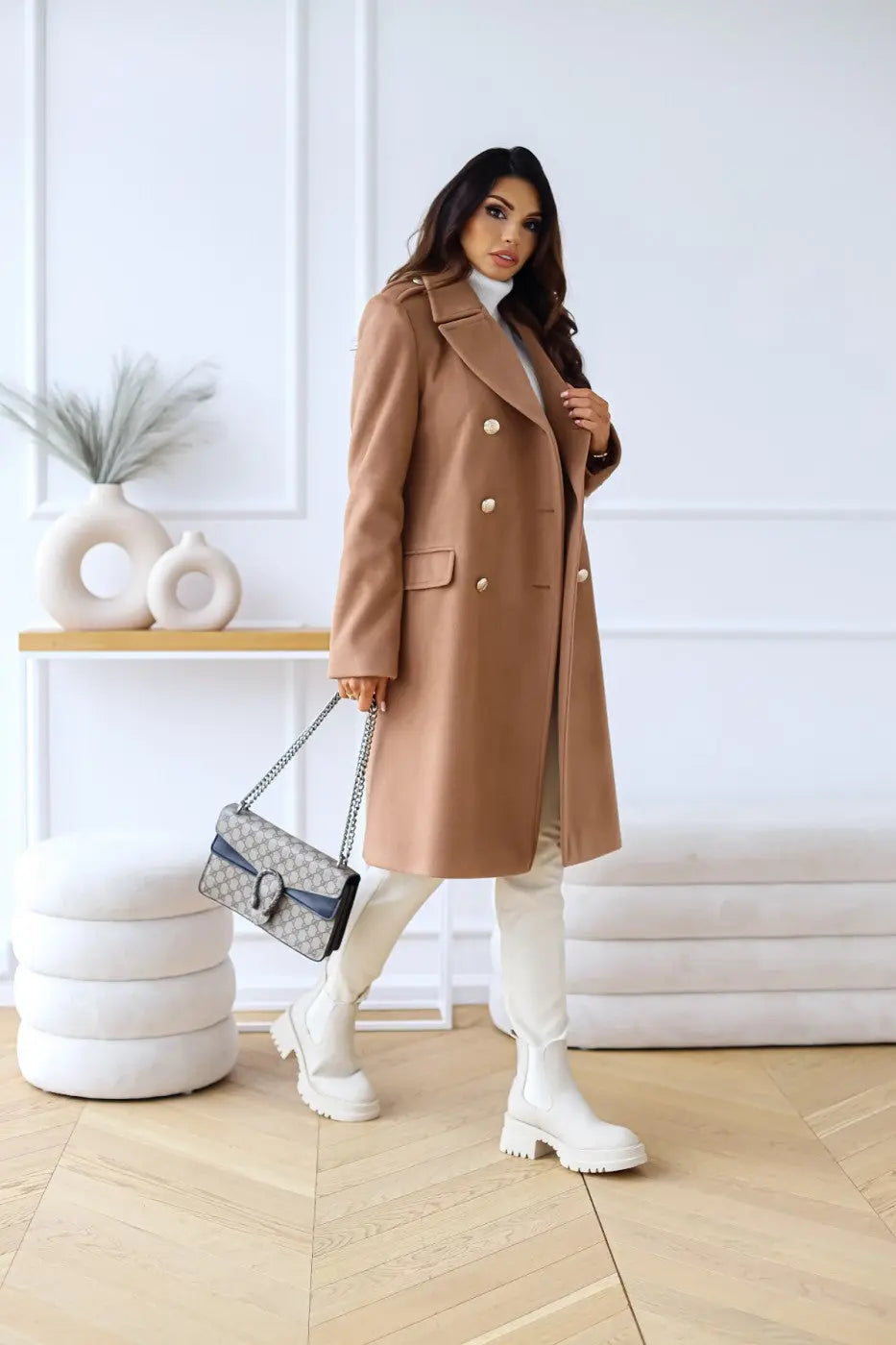 Kvinde i elegant brun frakke med hvide støvler og designerhåndtaske, står i moderne indretning. Mode, vintertøj, stilfuldt look.