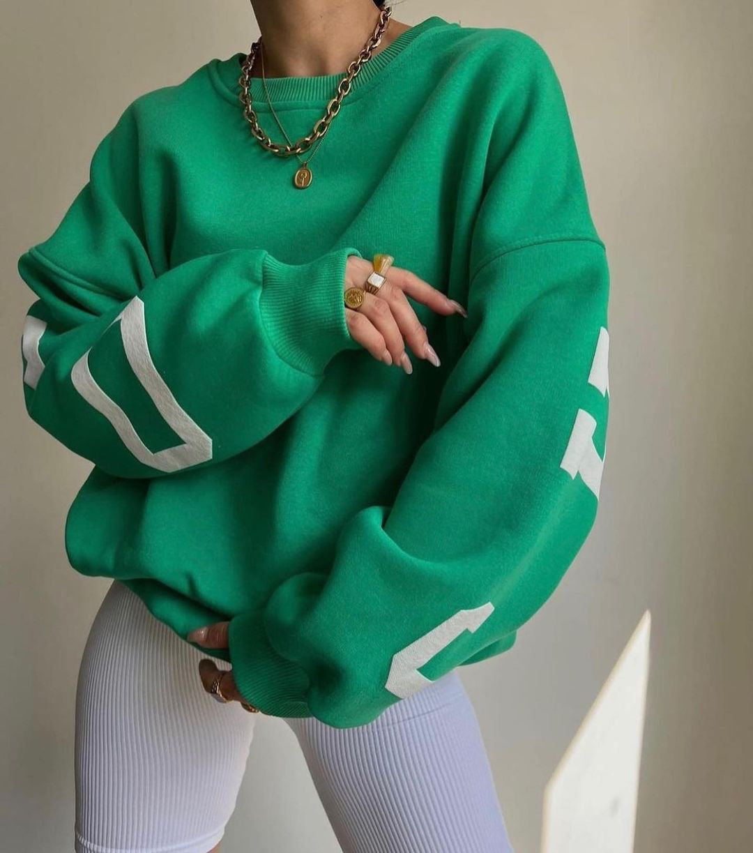 Kvinde iført grøn oversized sweatshirt med hvide tal, guldhalskæde og ringe. Moderne streetwear mode, trendy afslappet stil.