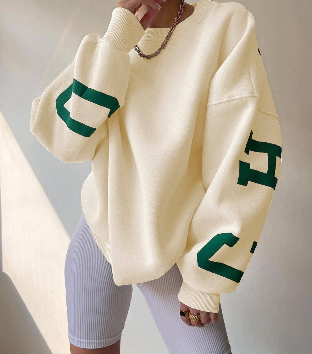 Beige oversized sweatshirt med grønne bogstaver, båret af person i hvide cykelshorts. Trendy streetwear mode, afslappet stil, moderne design.