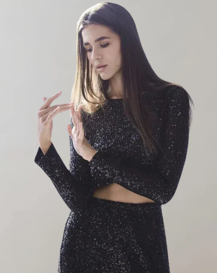 Ung kvinde i sort, glitrende kjole, ser ned på sine hænder. Langt, brunt hår, minimalistisk stil. Elegant mode, moderne kvindetøj, stilfuldt look.