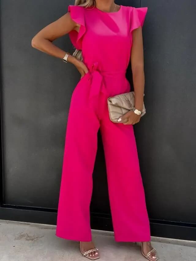 Kvinde i pink buksedragt med flæseærmer, holder beige clutch. Elegant mode, sommeroutfit, stilfuldt look. Perfekt til fest eller aftenbegivenhed.