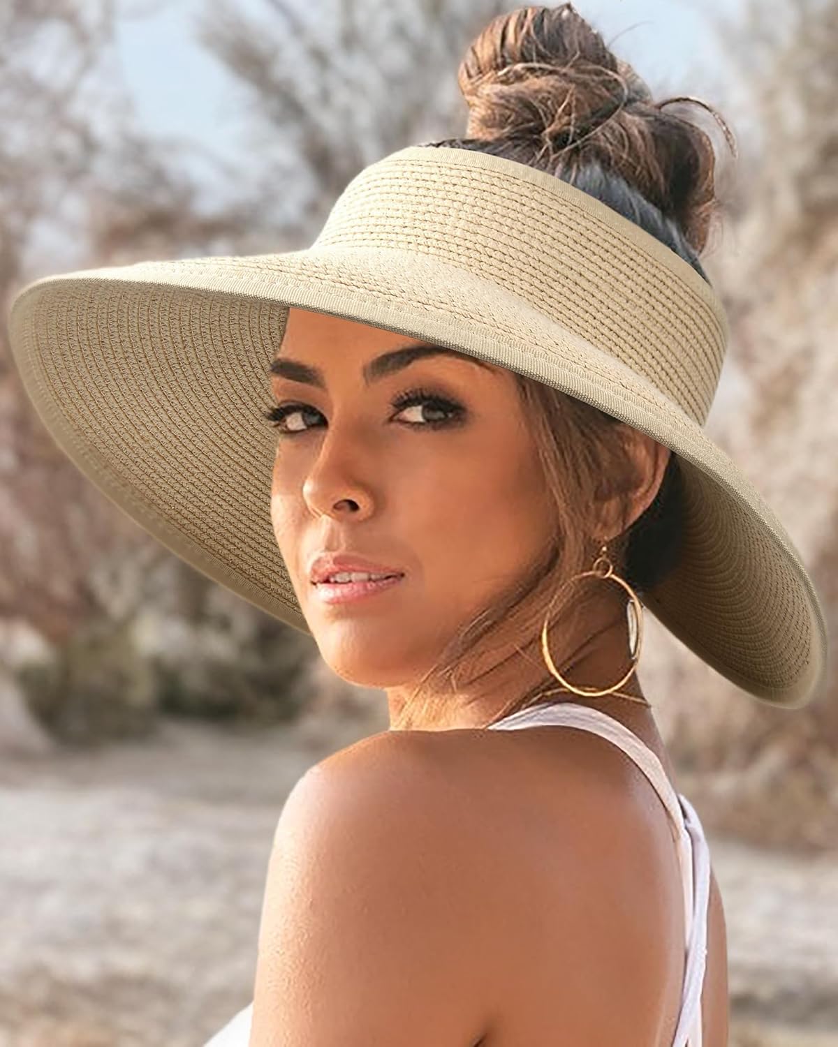 Kvinde med bredskygget solhat, gyldne øreringe og hvid top i naturligt udendørsmiljø. Sommermode, solbeskyttelse, stilfuldt tilbehør.