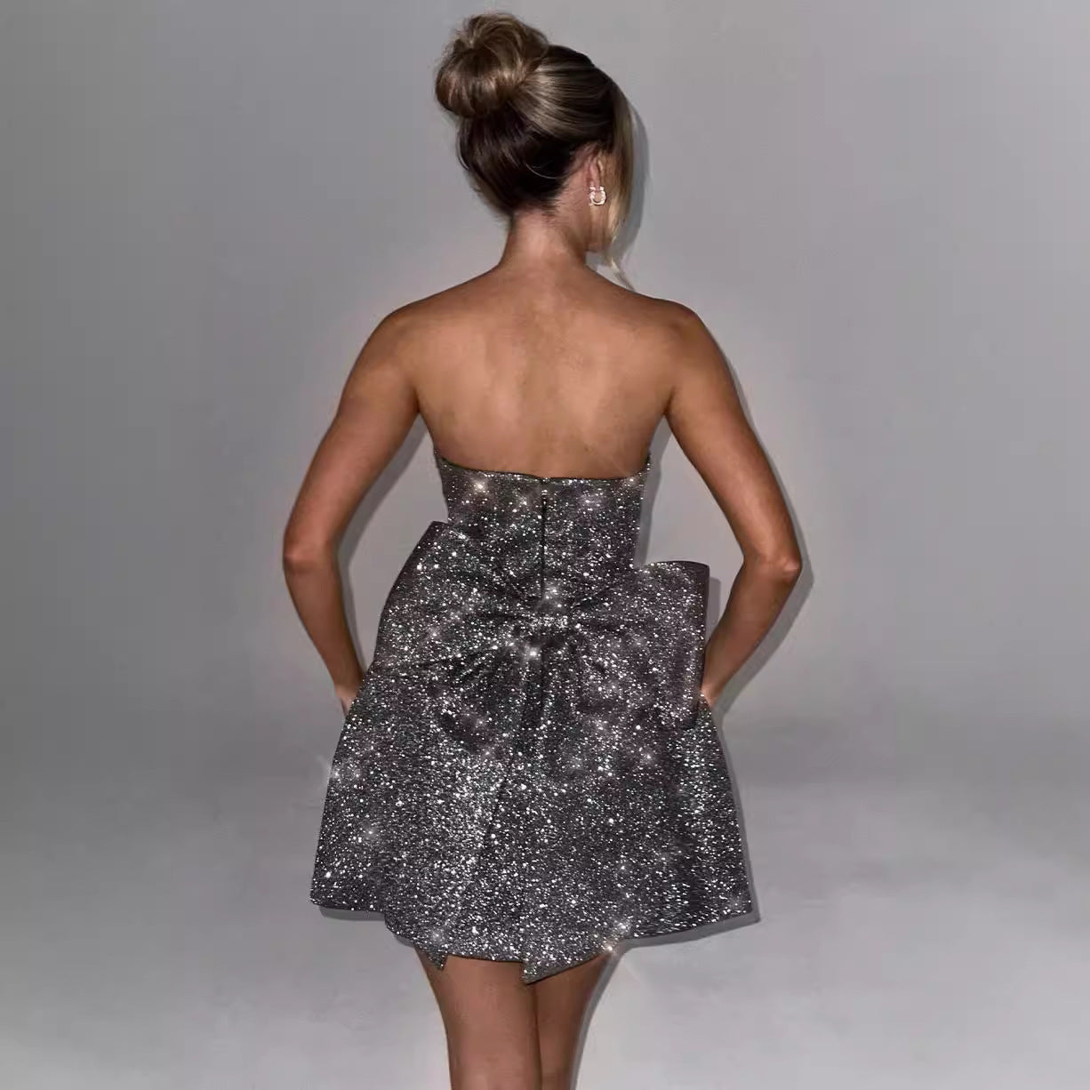 Kvinde i glitrende sølvkjole med stor sløjfe, set bagfra. Elegant aftenmode, opstrammet hår, minimalistisk baggrund.