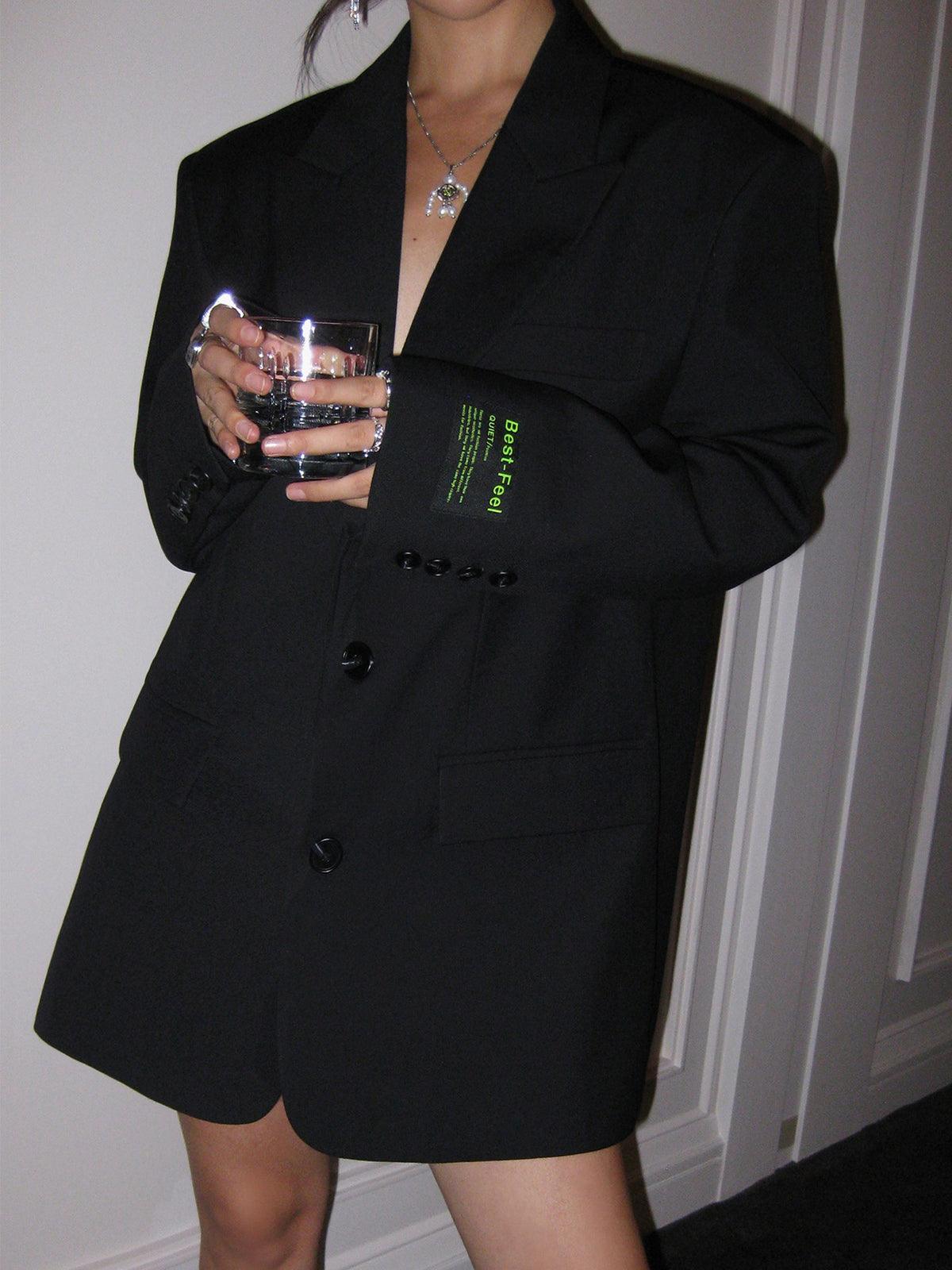 Kvinde i oversize sort blazer fra Balenciaga, holder et glas vand. Elegant mode, minimalistisk stil, luksusmærke, moderne kvindetøj.