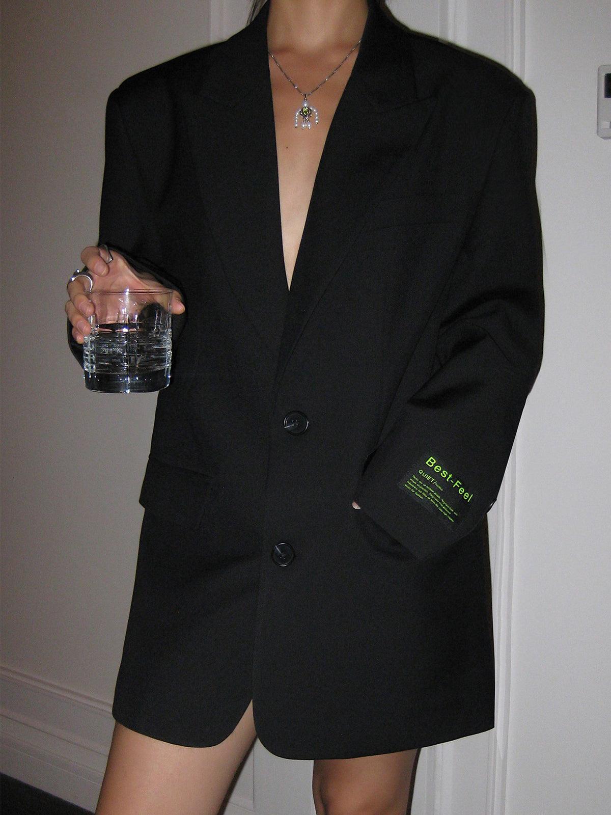 Kvinde i sort blazer med glas vand, minimalistisk mode, moderne stil, elegant outfit, trendy accessories, stilfuld halskæde, tidløs elegance.
