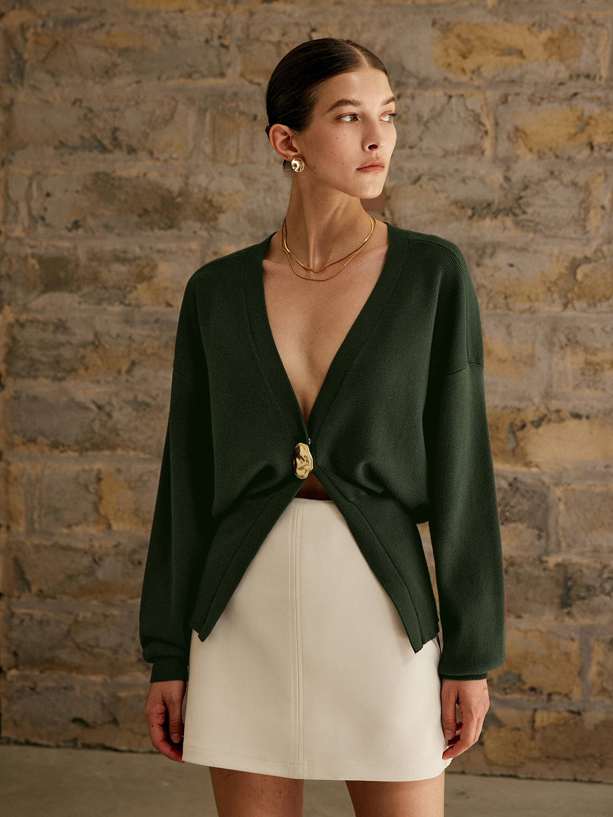 Kvinde iført mørkegrøn cardigan med dyb V-udskæring og hvid nederdel, står foran en murstensvæg. Elegant mode, minimalistisk stil, moderne kvindetøj.