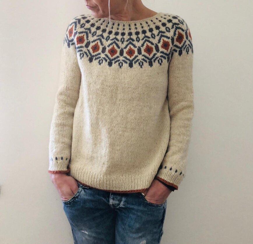 Beige striktrøje med nordisk mønster, båret af person i afslappede jeans. Håndstrikket uldtrøje, traditionelt design, vintermode.