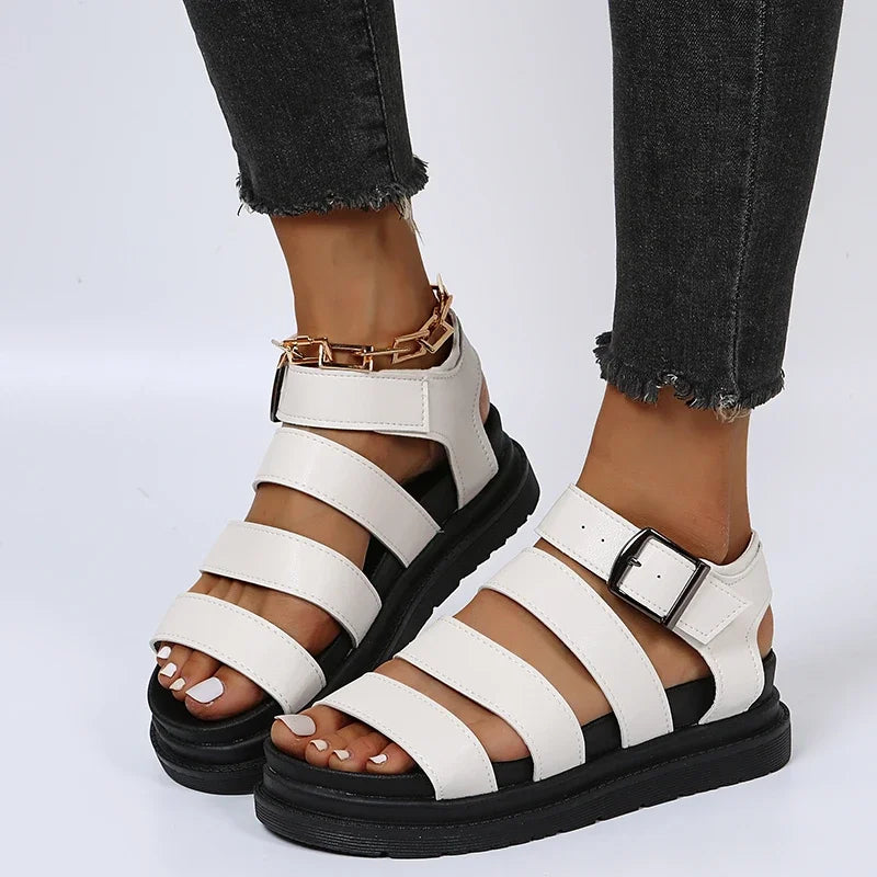 Dame iført hvide læder sandaler med sorte såler og guldkæde detalje, kombineret med sorte jeans. Moderne fodtøj, stilfuldt design.
