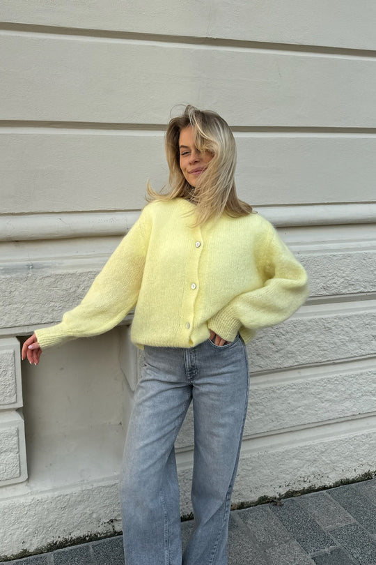 Kvinde iført gul striktrøje og grå jeans står foran hvid mur. Mode, efterårstøj, casual stil, trendy look, komfortabelt outfit.