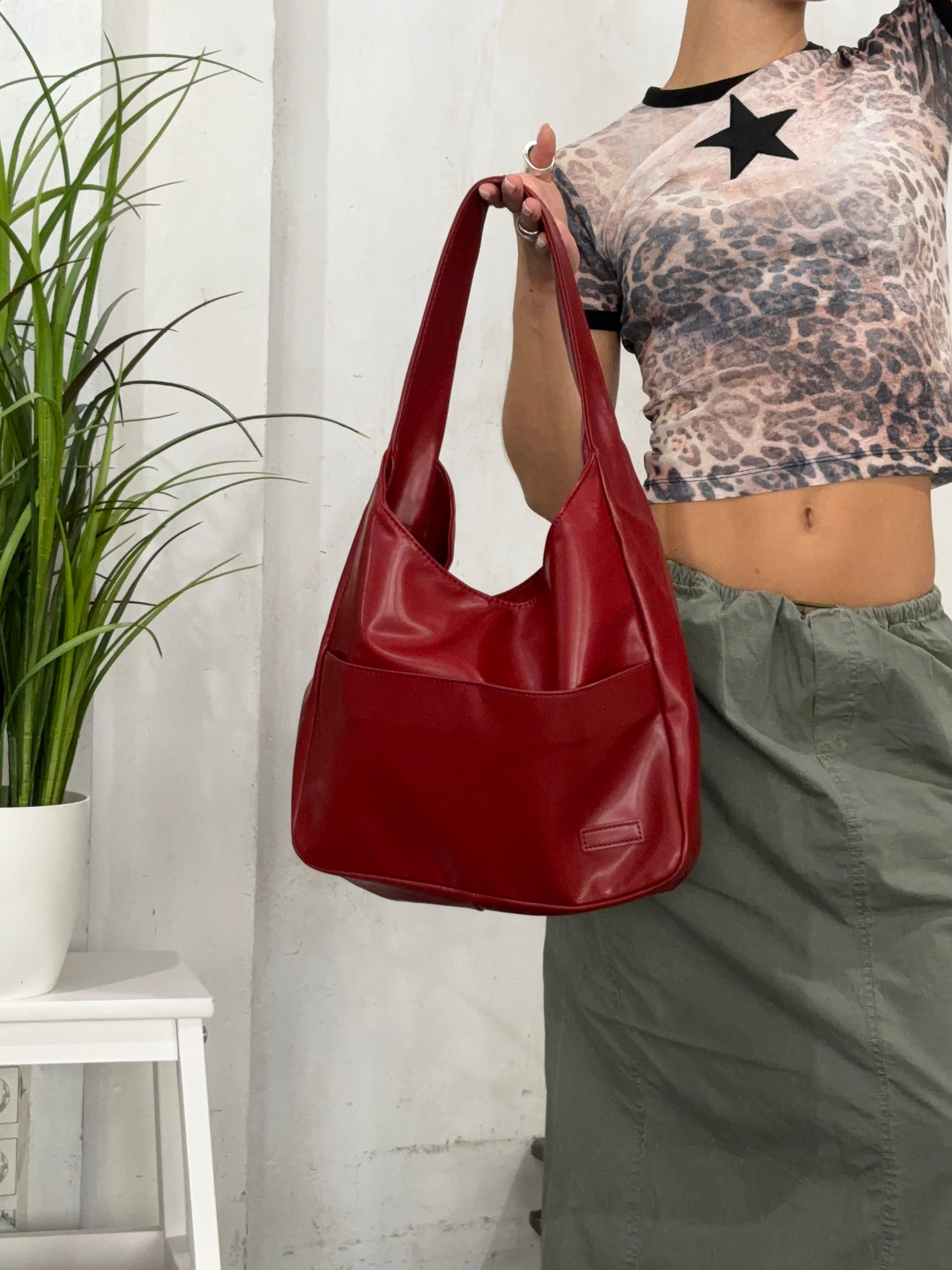Rød lædertaske holdt af person i leopardmønstret top og grøn nederdel. Moderne håndtaske, stilfuldt tilbehør, mode. Indendørs plante i baggrunden.