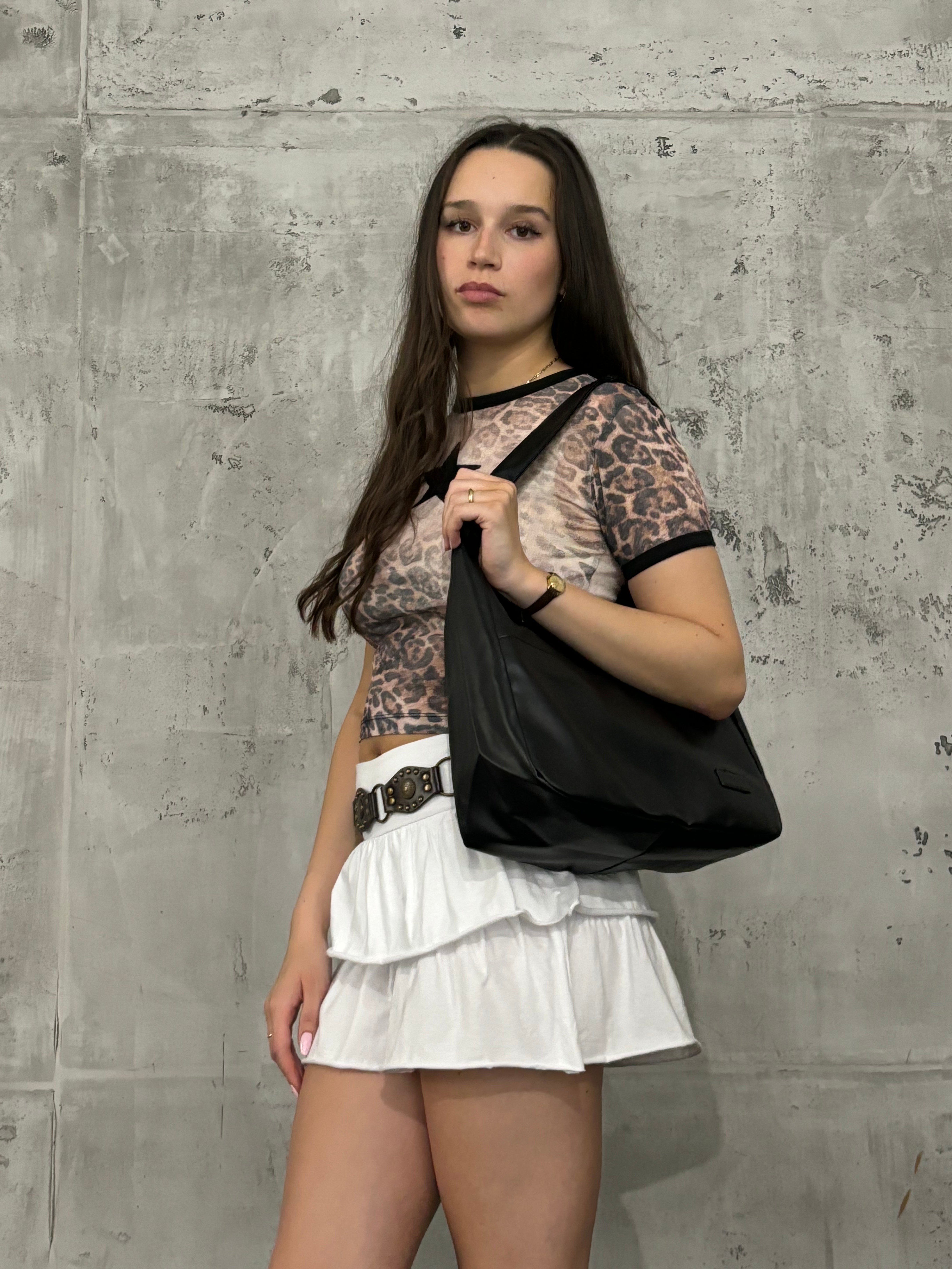 Ung kvinde i leopardmønstret top og hvid nederdel med sort skuldertaske foran betonvæg. Mode, trendy outfit, stilfuldt look.