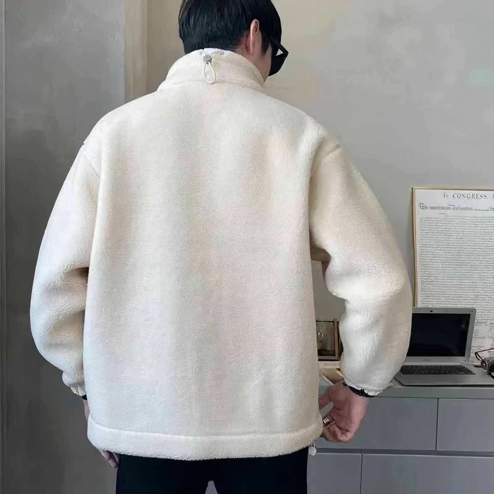 Mand iført hvid fleecejakke set bagfra, står ved skrivebord med bærbar computer og dokumenter. Moderne indretning, casual vintermode.