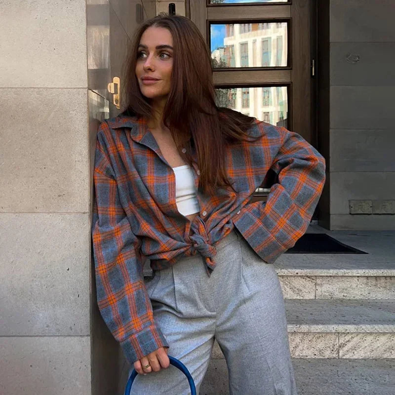 Ung kvinde i ternet skjorte og grå bukser står afslappet op ad en mur. Mode, street style, moderne kvindetøj, afslappet look.