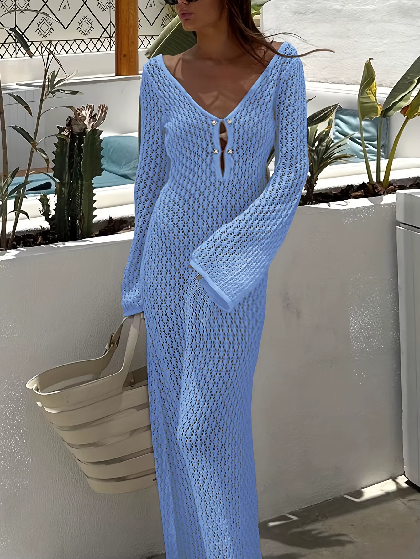 Kvindelig model i lyseblå, hæklet maxikjole med dyb V-udskæring og lange ærmer, poserer udendørs ved hvid mur med planter. Sommermode, elegant stil.