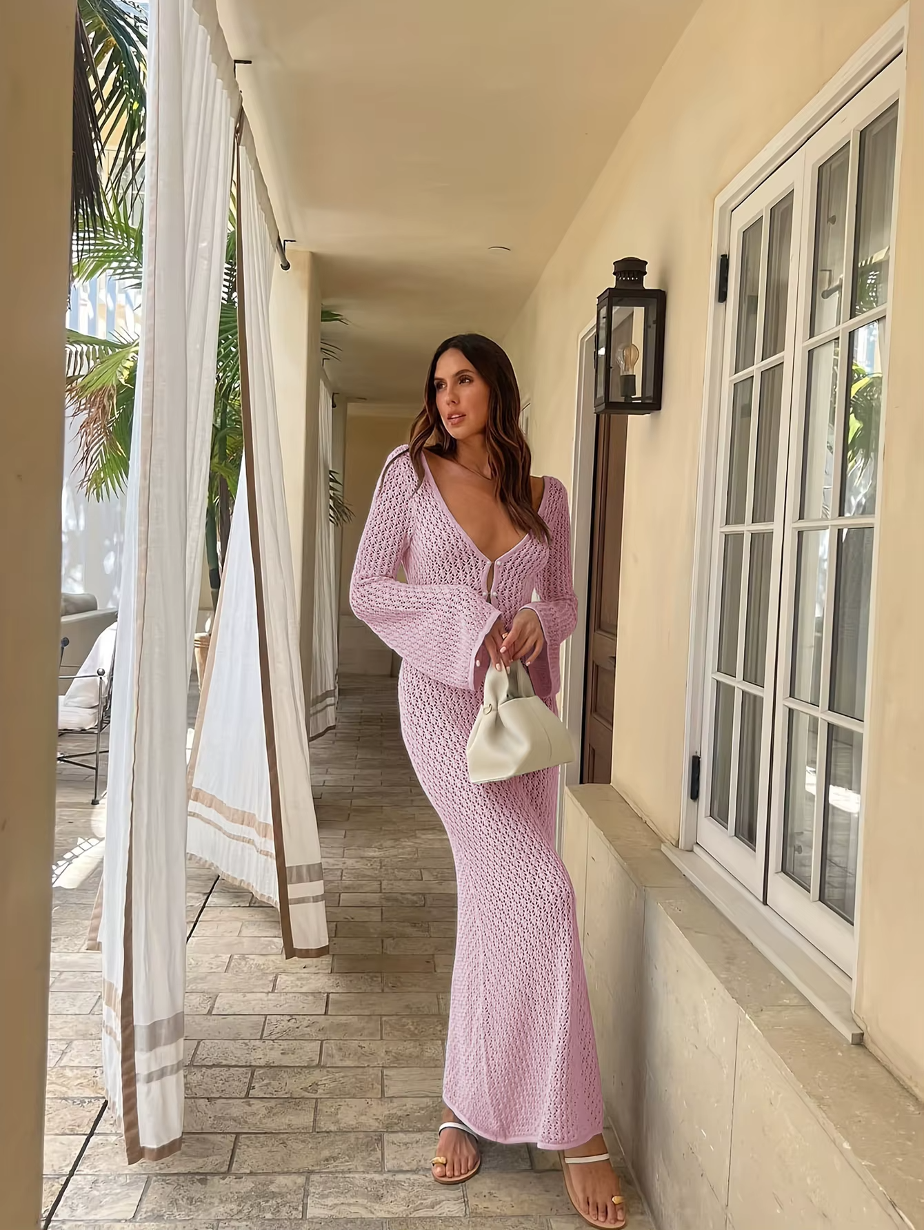 Kvinde i lang, lyserød strikkjole med dyb V-udskæring, poserer på terrasse. Hun holder en hvid taske. Sommermode, elegant stil, udendørs.