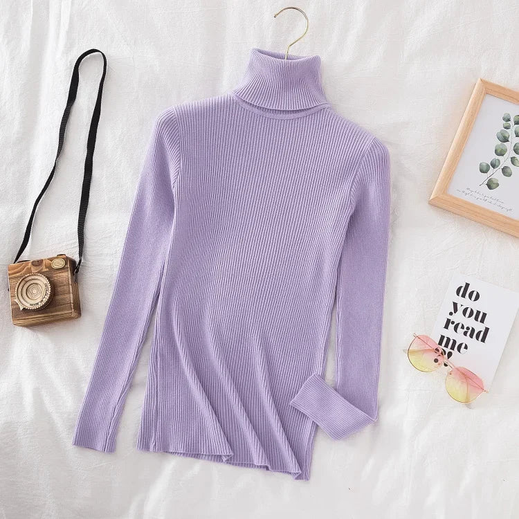 Lyselilla rullekrave sweater i ribstrik på hvid baggrund, ved siden af retro kamera, solbriller og indrammet plantebillede.