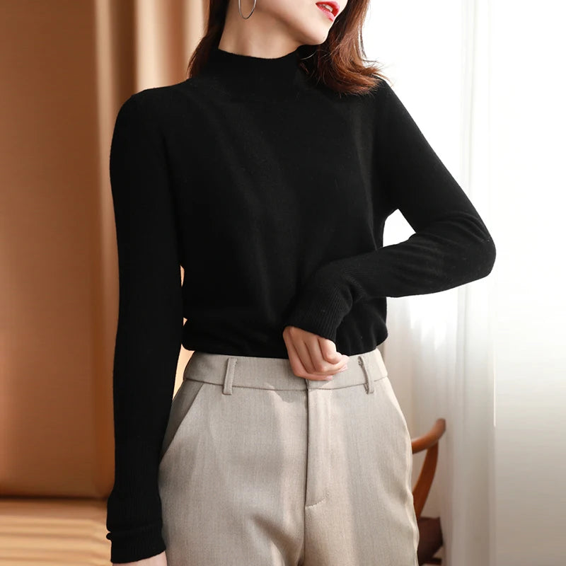 Kvinde iført sort sweater og beige bukser, står ved vindue. Elegant mode, minimalistisk stil, efterårstøj, moderne kvindemode, skandinavisk design.