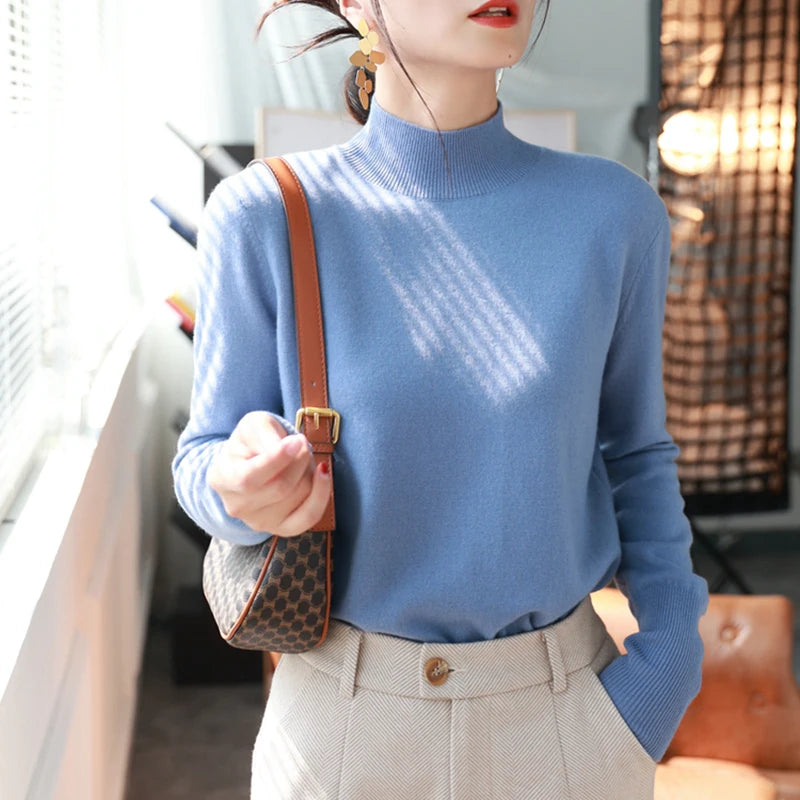 Kvinde i blå sweater og beige bukser, bærer en brun skuldertaske. Moderne mode, elegant stil, indendørs belysning.