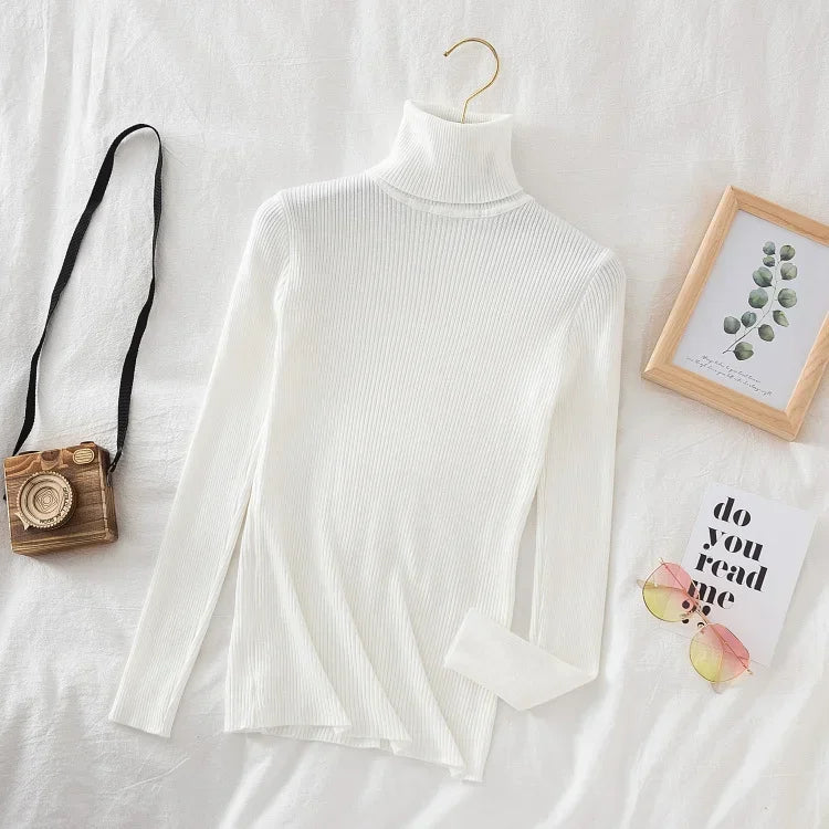 Hvid rullekrave sweater på seng, retro kamera, briller og plantebillede. Moderne minimalistisk mode, stilfuldt tøj, skandinavisk design.