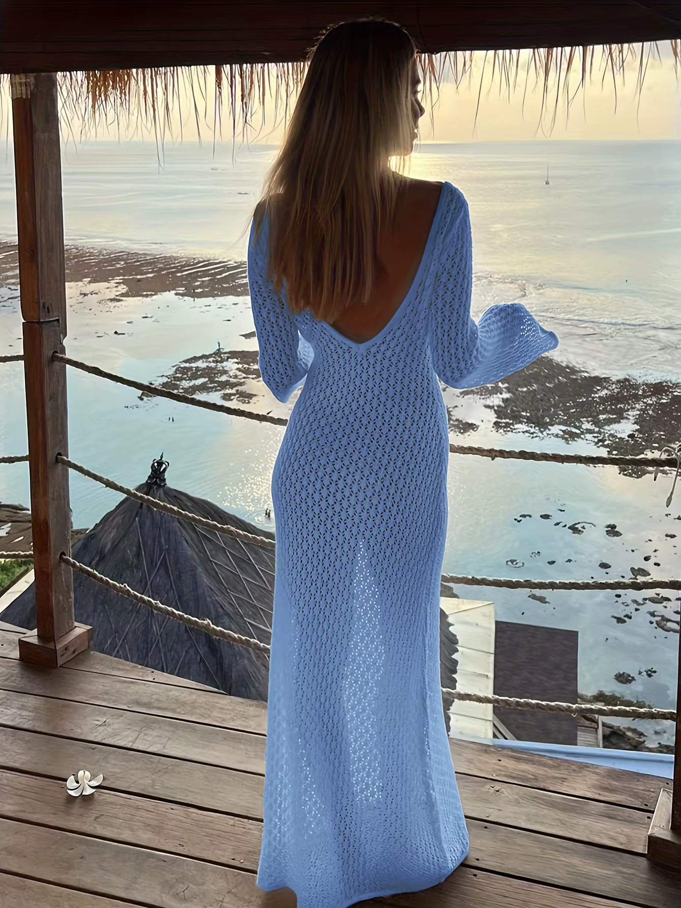 Kvinde i blå, hæklet kjole med dyb rygudskæring står på terrasse med udsigt over havet ved solnedgang. Strand, havudsigt, sommermode.