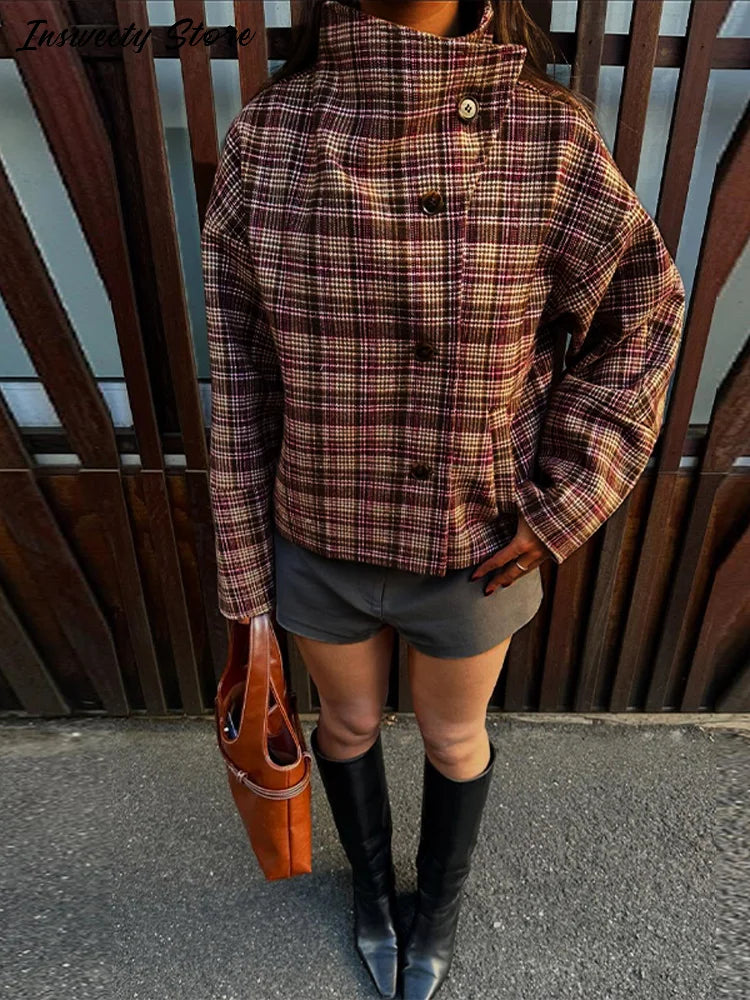 Kvinde iført ternet jakke, grå nederdel og sorte støvler, holder brun taske foran træport. Mode, efterårstøj, stilfuldt look.