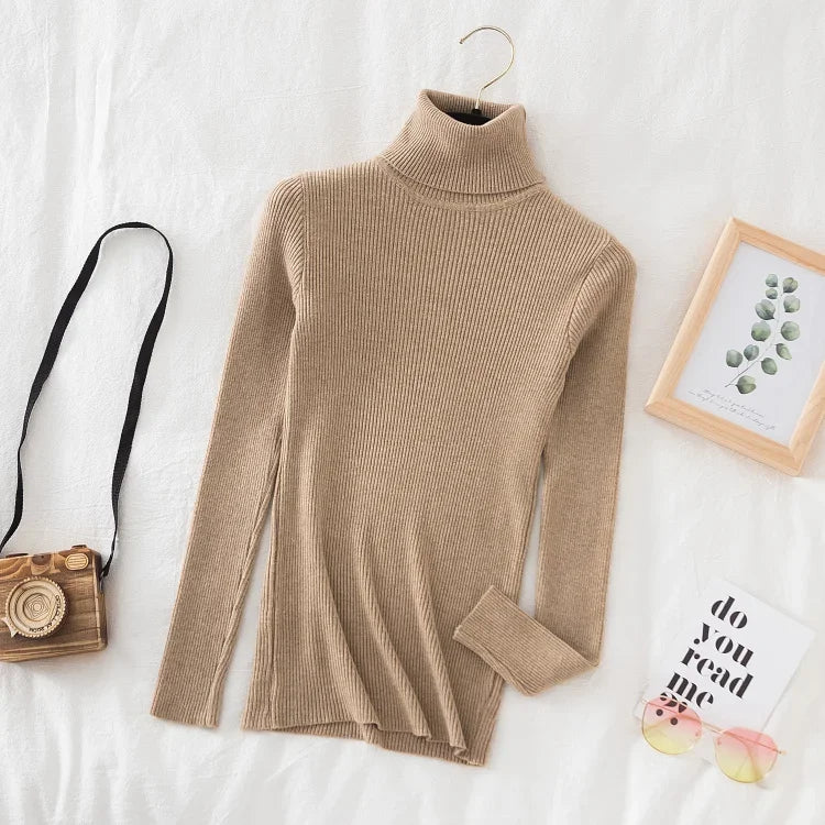 Beige rullekrave sweater på hvidt sengetøj, ved siden af et kamera, en ramme med planteillustration og en bog. Stilfuldt og minimalistisk design.