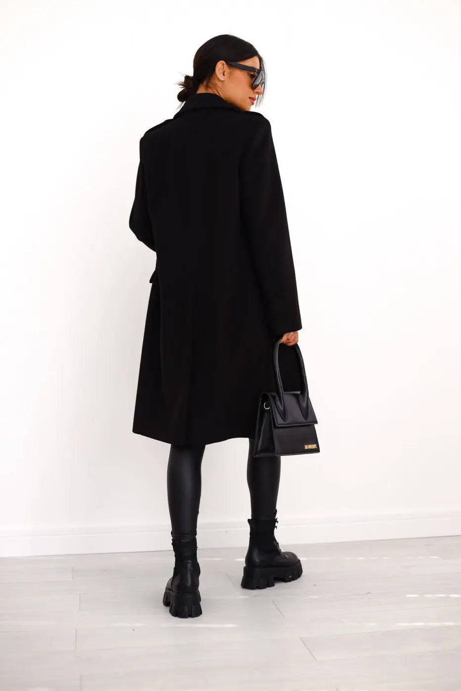 Kvinde i sort frakke og solbriller, bærer sort håndtaske og støvler, står mod hvid baggrund. Elegant mode, minimalistisk stil, vintertøj.