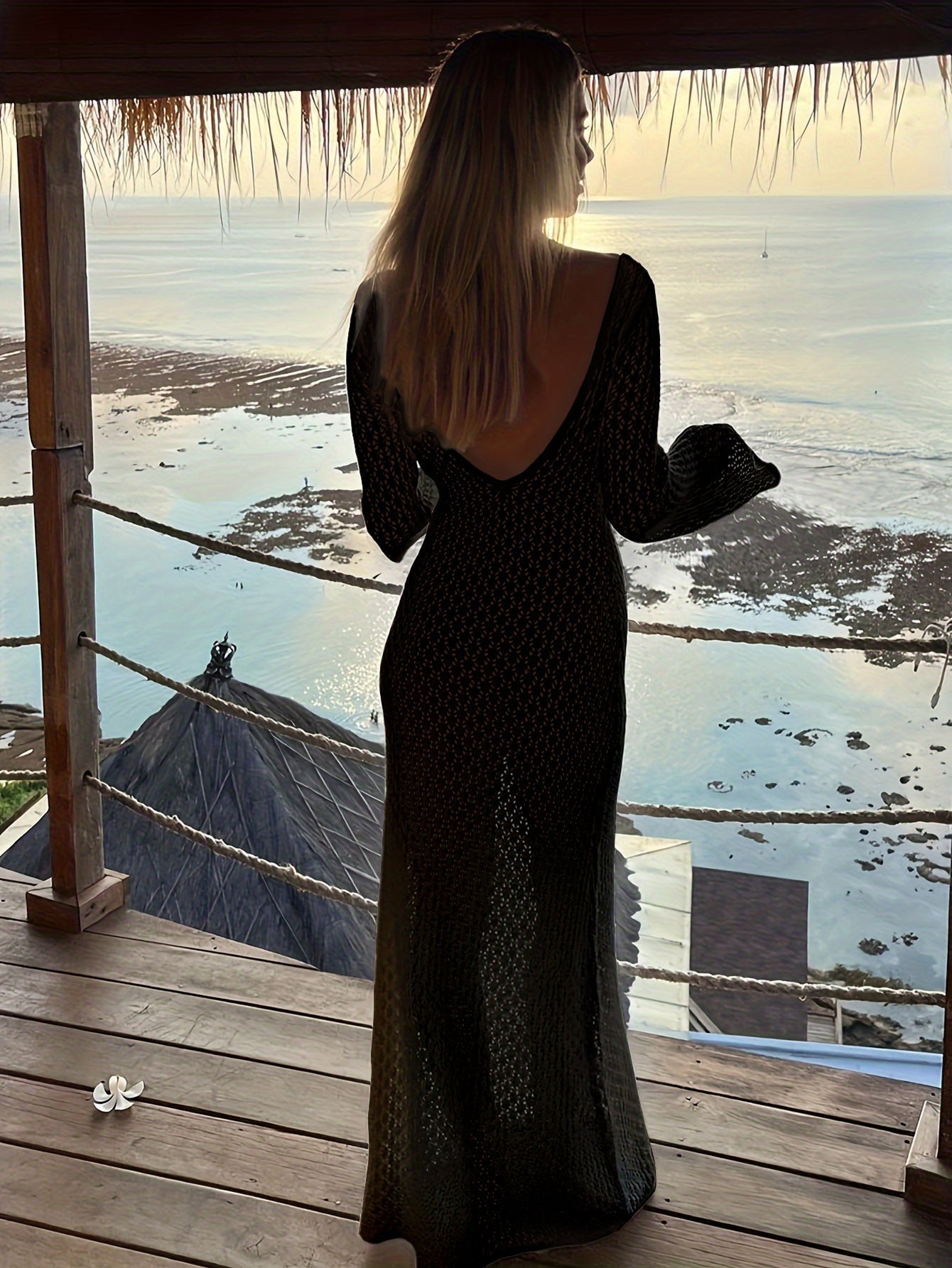 Kvinde i sort kjole står på balkon med udsigt over havet ved solnedgang. Strand, havudsigt, solnedgang, ferie, romantisk atmosfære.