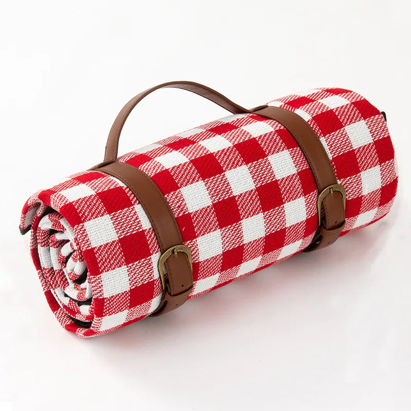Rød og hvid ternet picnic-tæppe med læderremme, sammenrullet til transport. Perfekt til udendørs brug, camping og strandture.